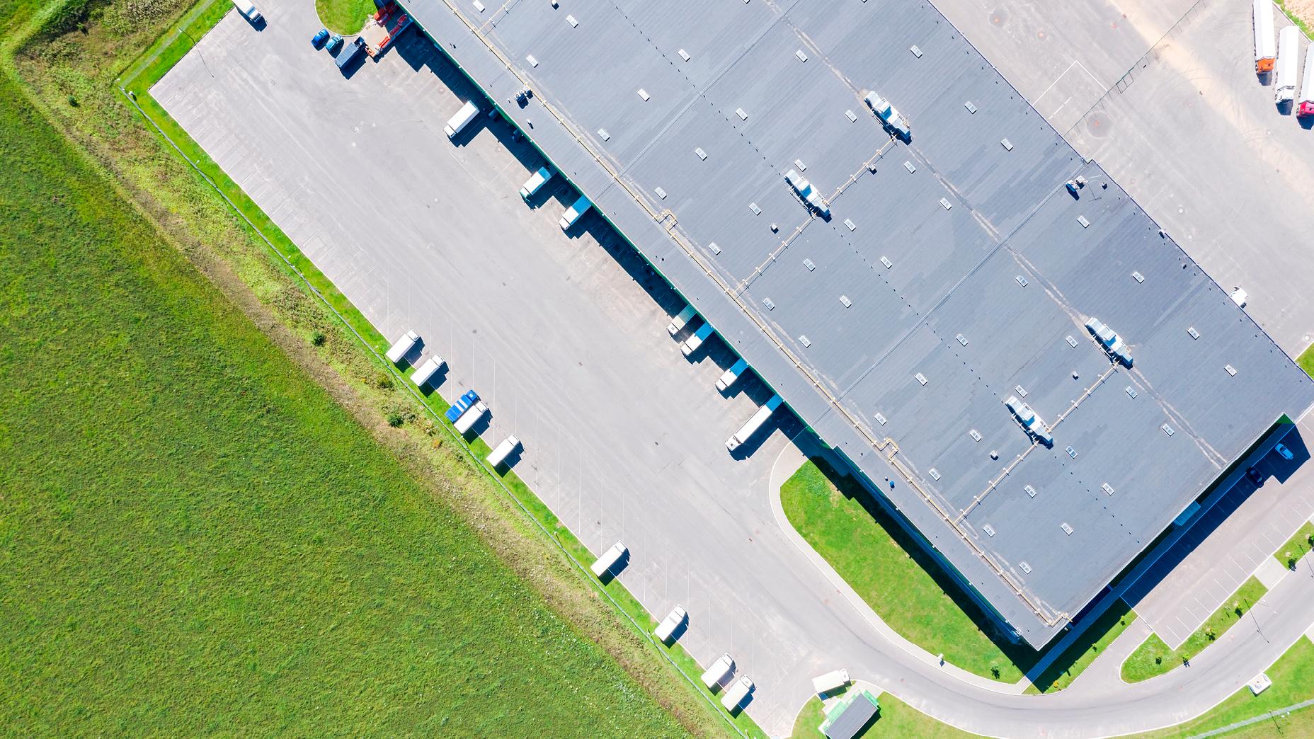 Aerial photograph of a large warehouse with multiple loading bays and parked trucks, surrounded by grass fields.