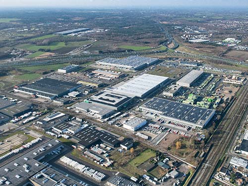 Aerial view of an industrial area with large warehouses, roads, railway lines and in the background green fields and residential areas under a partly cloudy sky.