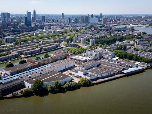 Aerial view of a city with a river in the foreground, surrounded by residential and office buildings, green areas and a peninsula of industrial buildings.