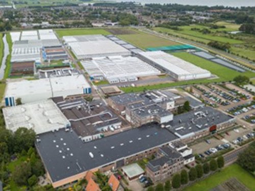 Aerial view of a large industrial complex with several buildings, surrounded by farmland, greenhouses and some residential houses.