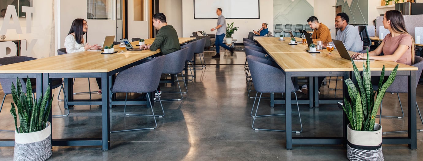 Open-plan coworking space with long shared desks, people working on laptops and a lounge area in the background.
