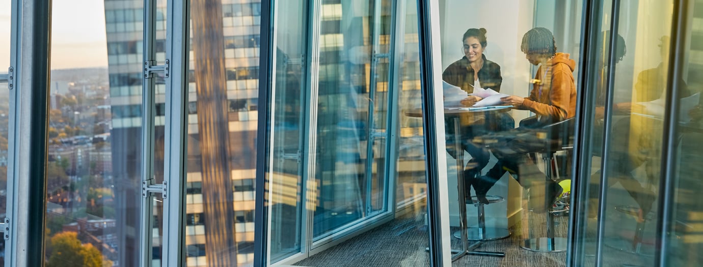 Two colleagues review documents in a glass-walled meeting room on a high floor of a London office tower.