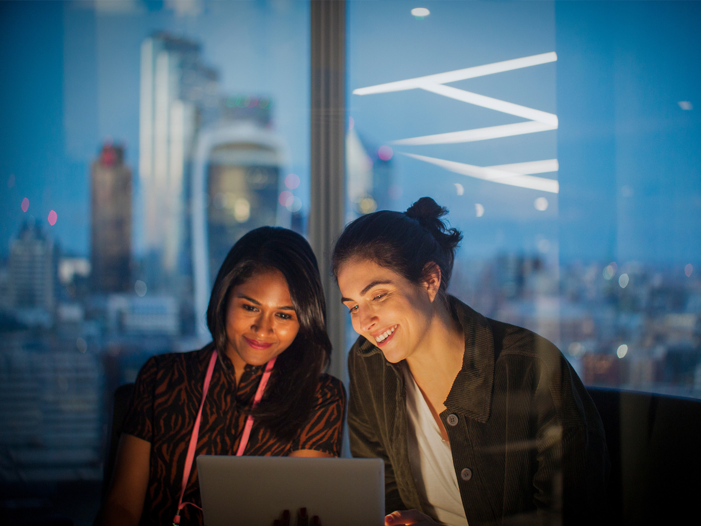 Two colleagues review work on a laptop beside floor-to-ceiling windows overlooking the Thames and the City of London skyline.
