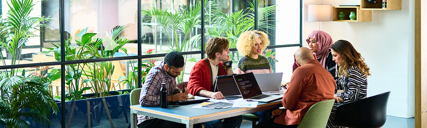 A group of people collaborate around a table with laptops in a bright, modern office filled with lush green plants.