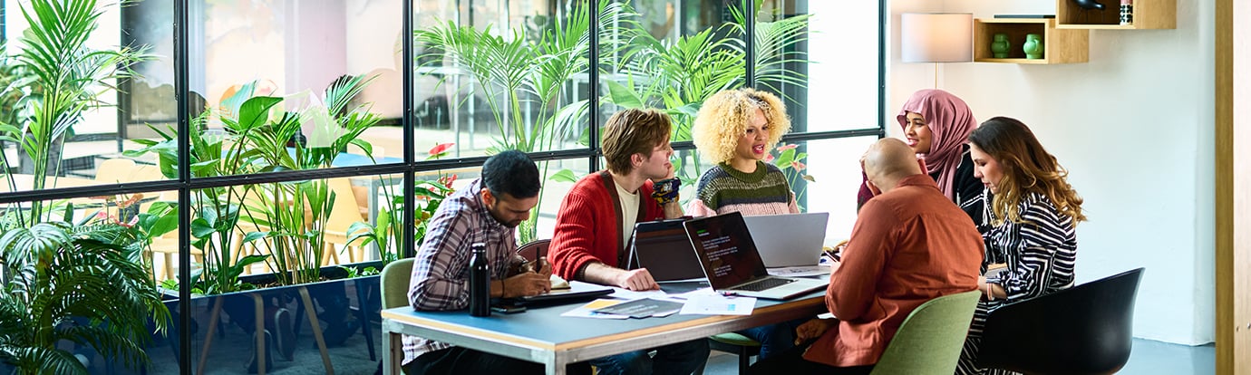 A group of people collaborate around a table with laptops in a bright, modern office filled with lush green plants.