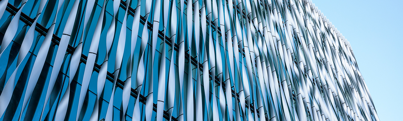 An architectural view of a modern glass building featuring a facade of vertical, wave-shaped white fins against a bright blue sky.