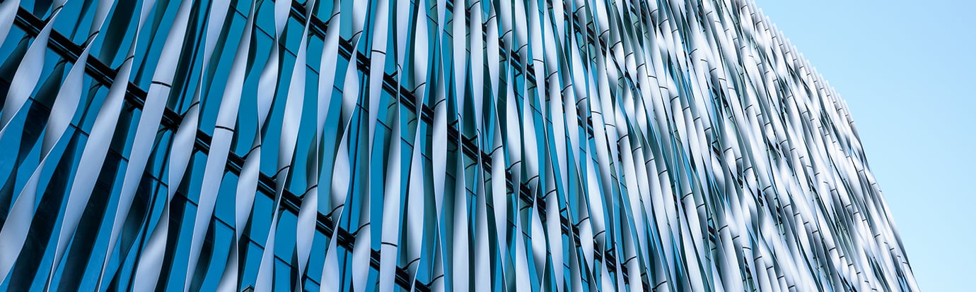 An architectural view of a modern glass building featuring a facade of vertical, wave-shaped white fins against a bright blue sky.