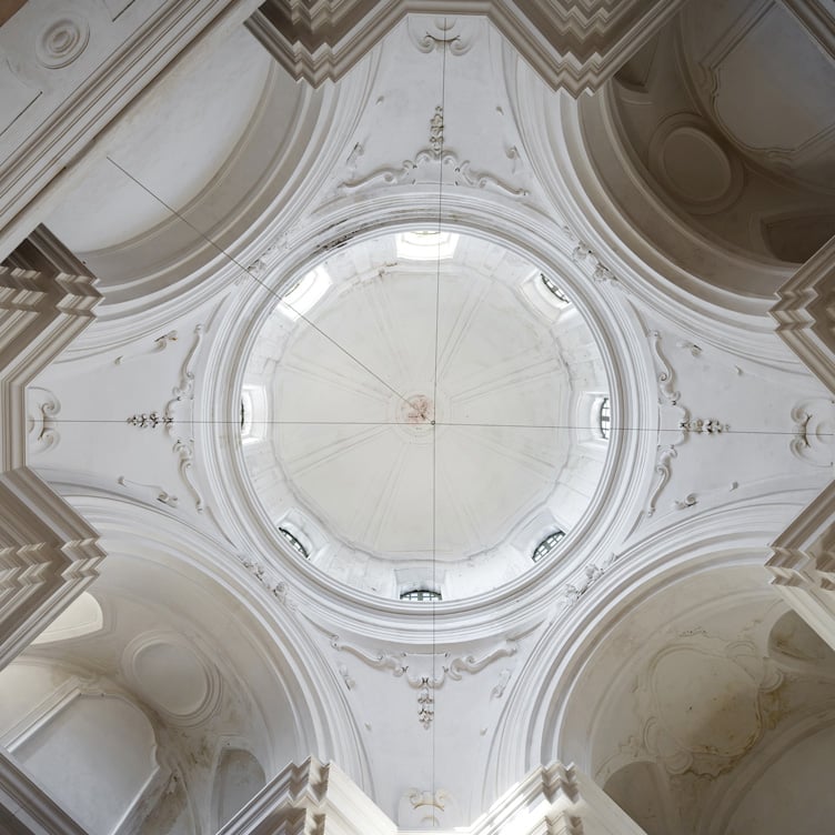A photograph looking up at the intricate dome of a Baroque-style building, featuring detailed white plasterwork, ornate molding, and arched windows allowing natural light to illuminate the elegant architectural design.