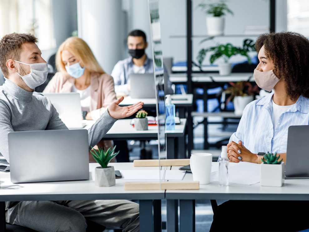 Young guy and african american woman in protective mask talking through glass board at workplace