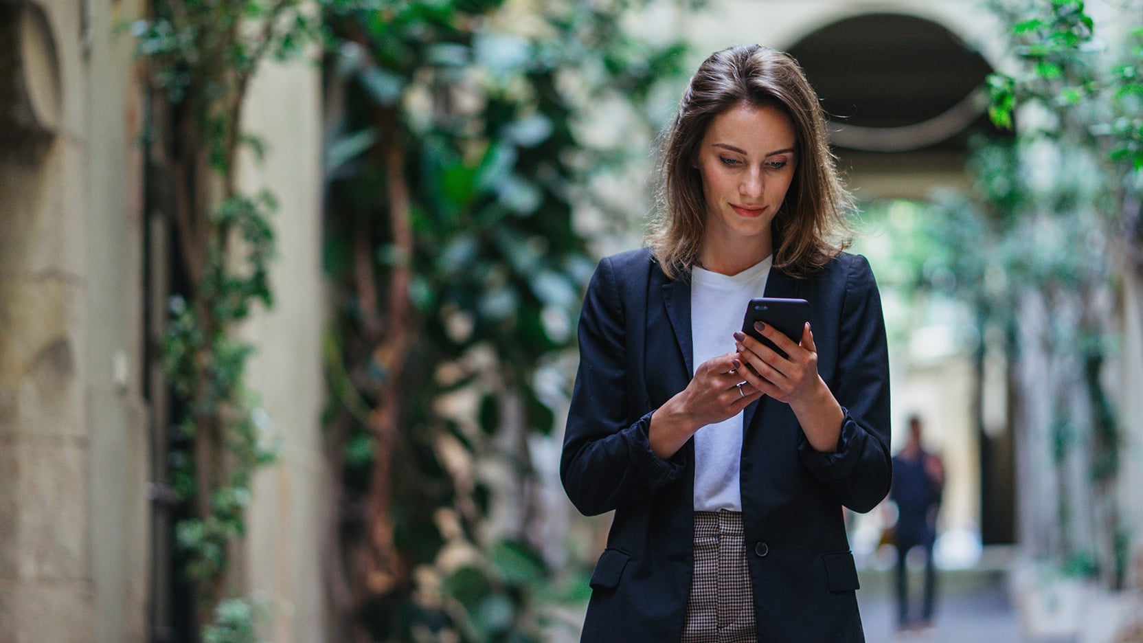 Professional woman in a navy blazer reading CBRE news and announcements on her smartphone while walking through a sunlit, ivy-covered courtyard.