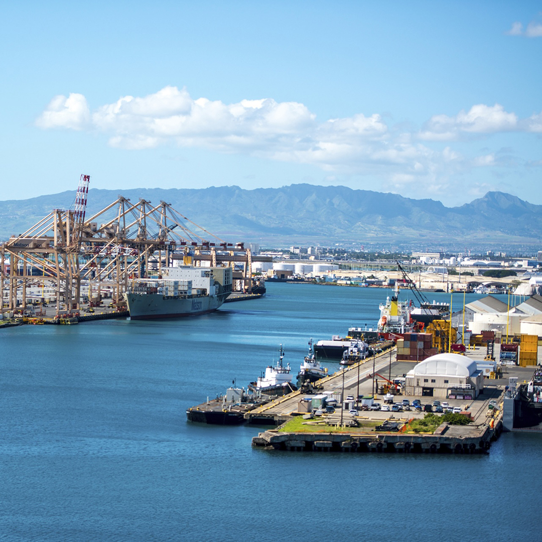 Aerial view of Honolulu Harbor showing container cranes, a Matson cargo ship at berth, tugboats, and industrial waterfront facilities with the Oahu mountain range in the background.