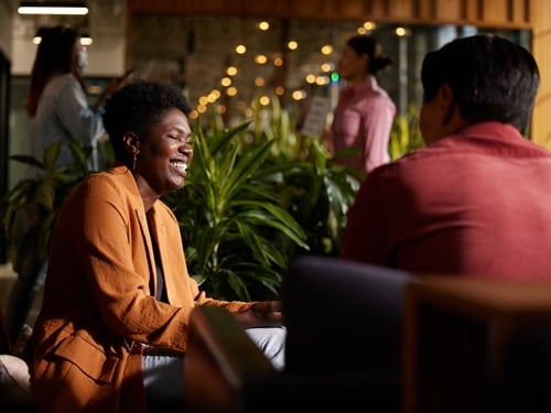 Two colleagues engaged in conversation in a warmly lit modern workplace lounge with greenery
