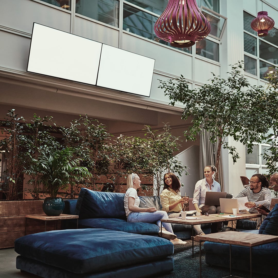 Group of professionals talking and working on laptops in a plant-filled office lounge with blue velvet seating, representing CBRE Workplace Strategy consulting that aligns space, technology, culture and operations to business performance.