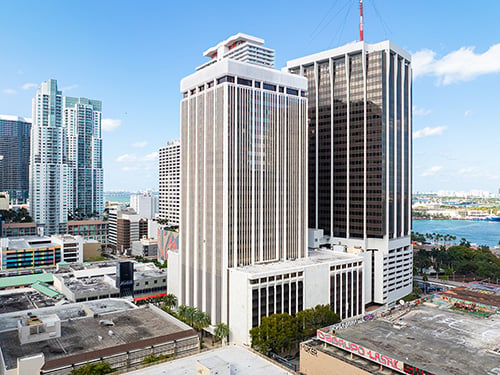Aerial view of One Downtown Miami office tower featuring a white vertical-fin high-rise facade with Biscayne Bay and Miami Beach visible in the background.