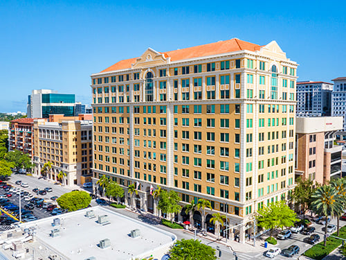 Aerial exterior of The Ponce, a Mediterranean Revival Class A office tower with terracotta roof, ornate cream facade, and palm tree-lined streetscape in South Florida.