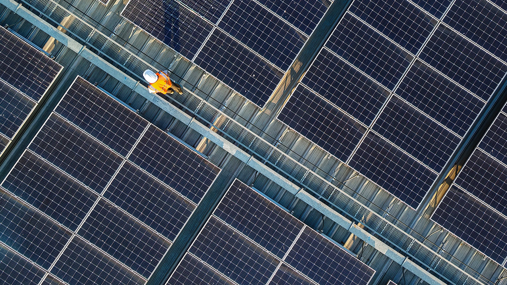Aerial view of rooftop solar panels with a worker in safety gear walking along a metal maintenance walkway.