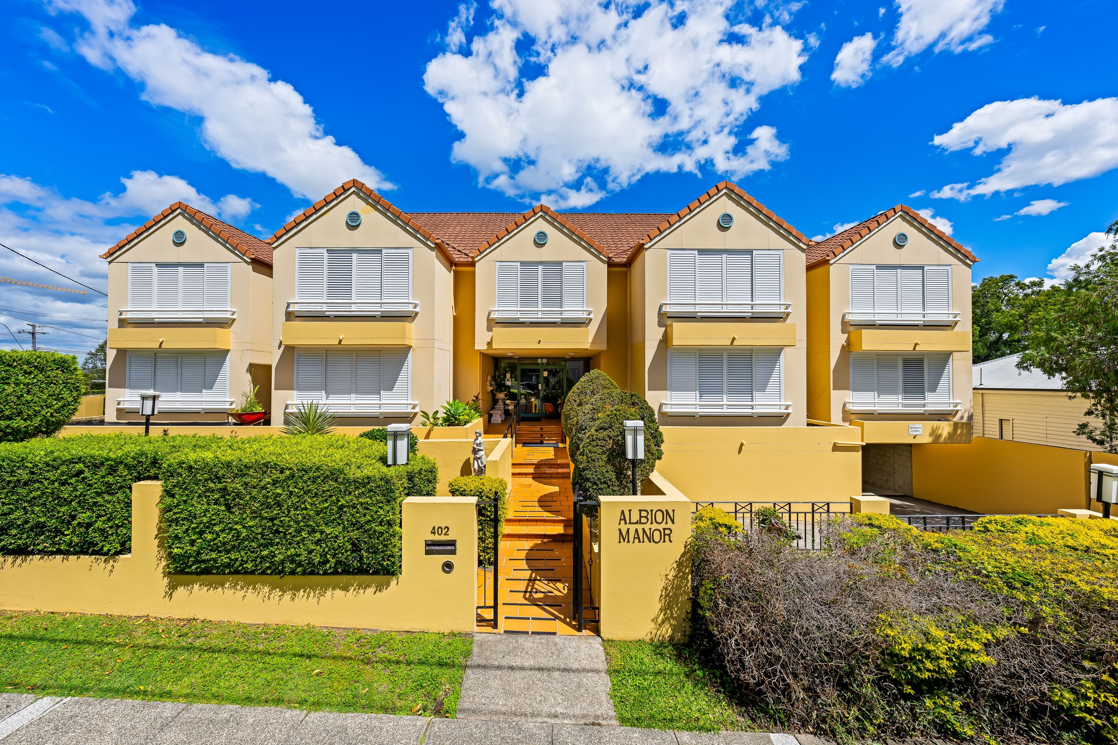 Albion Manor, a yellow two-storey apartment building with a tiled roof and white window shutters, set behind a manicured hedge under a bright blue sky.
