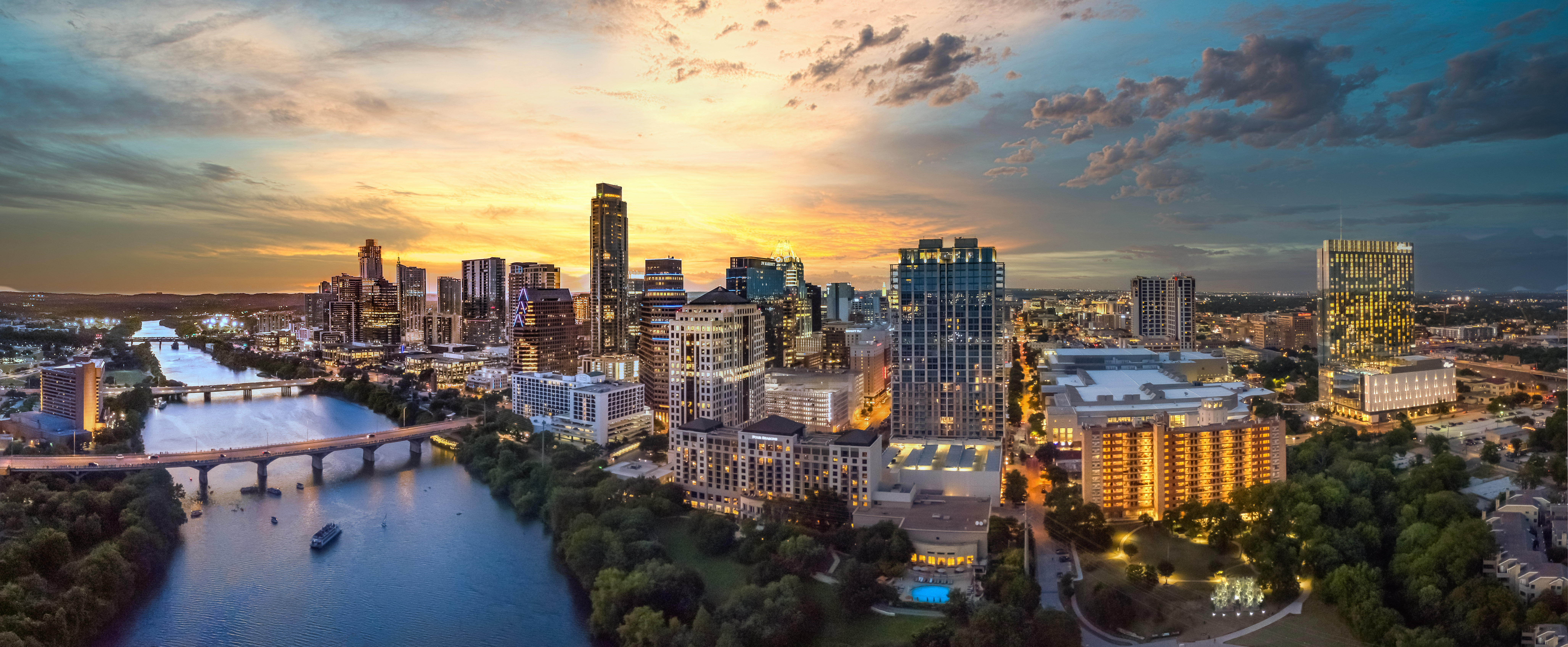 Downtown city skyline at sunset with high-rise commercial buildings along a river and illuminated bridges.