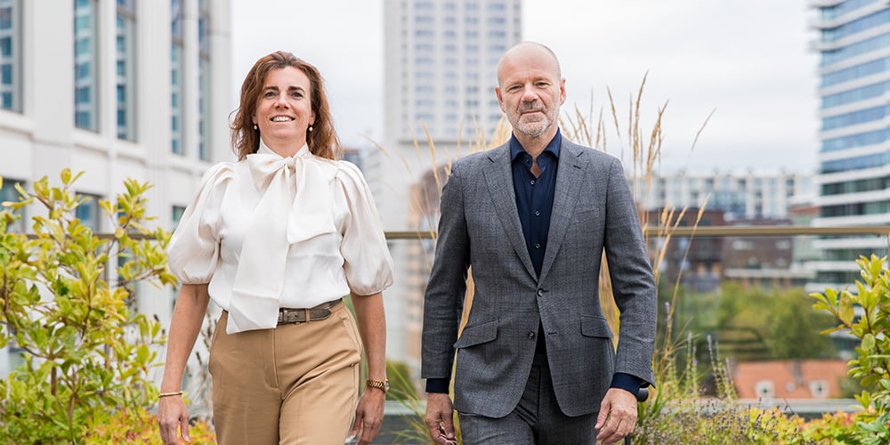 Two people walking towards the camera with a green environment between several tall office buildings in the background.