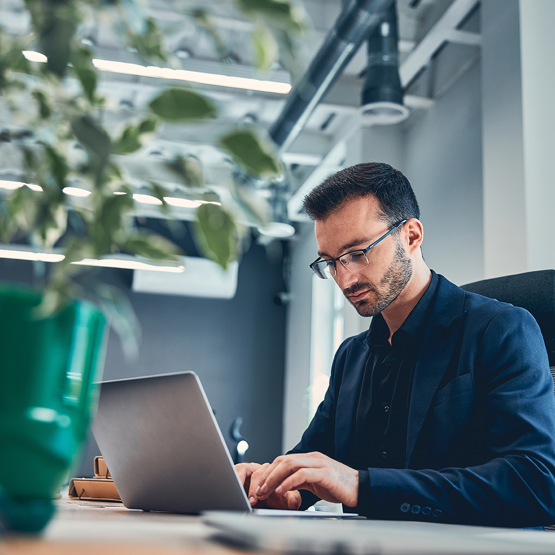 Business professional typing on a laptop in a modern office environment with natural light and greenery