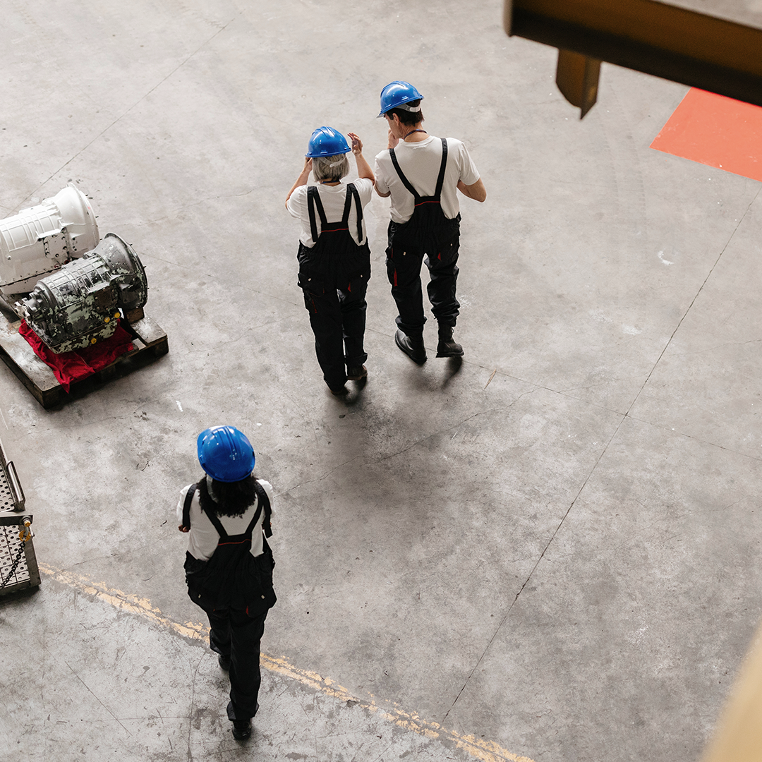 Three workers in blue hard hats and overalls walk across a factory floor, seen from above