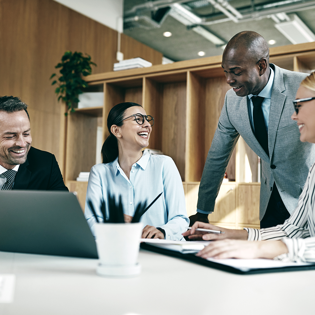 Four diverse business professionals laughing and collaborating around a conference table with a laptop in a modern office setting.
