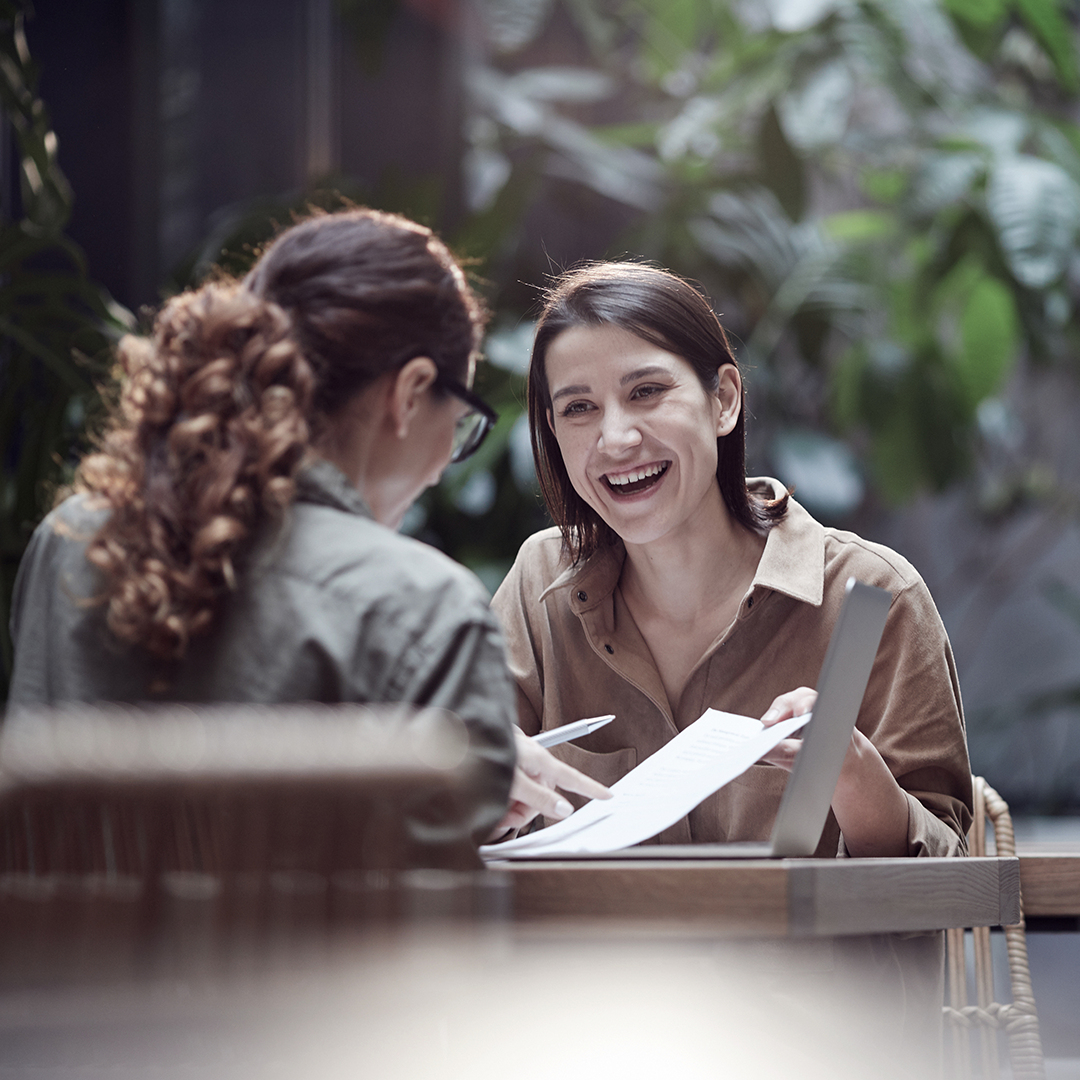 Two women smile and review documents together at a table with a laptop and greenery behind