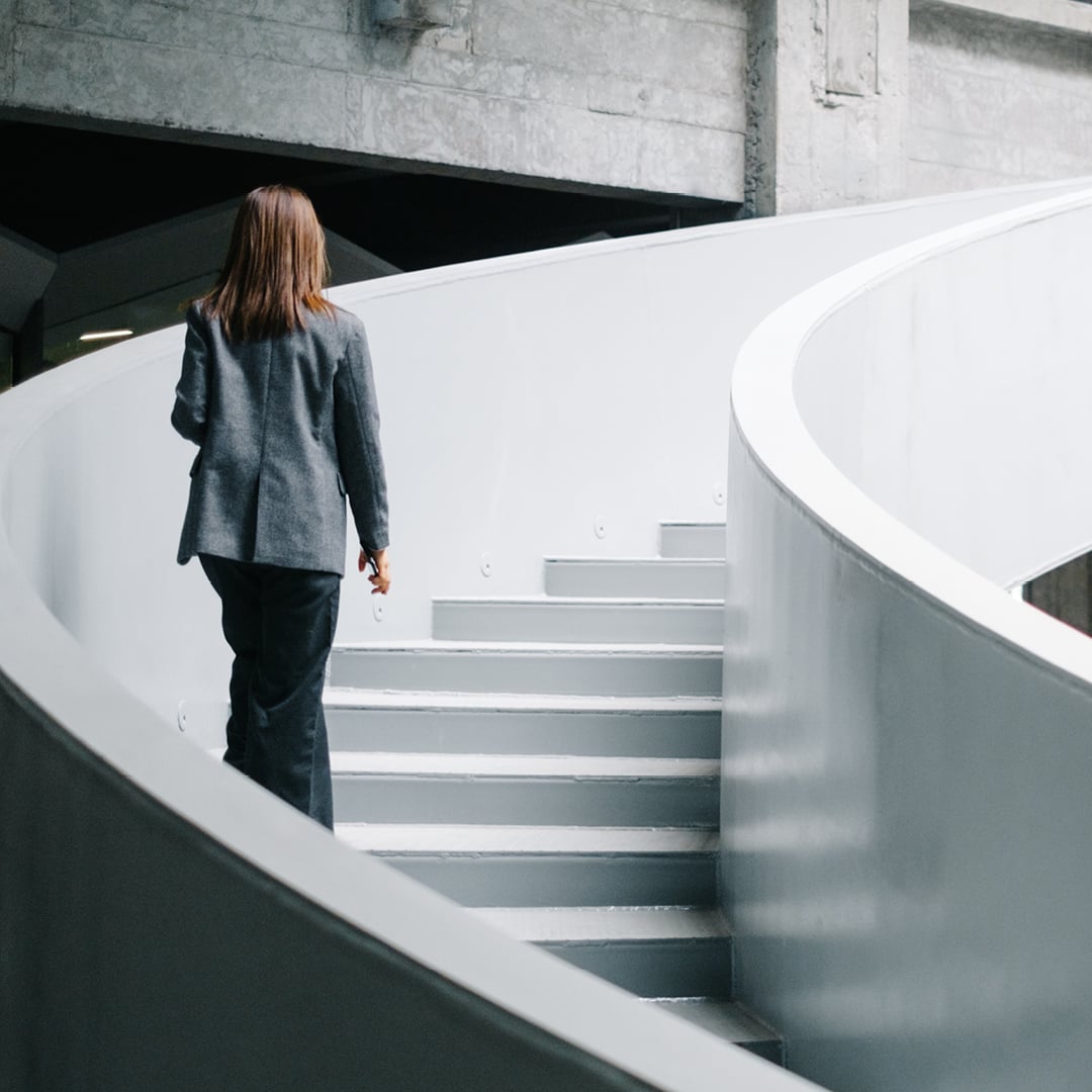 A businesswoman in a gray blazer ascends a curved white staircase in a modern commercial building symbolizing career advancement and workplace leadership.