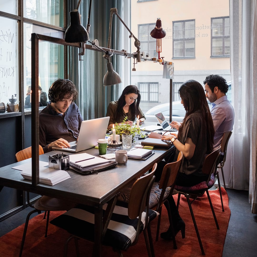 Group collaborating around a shared table in a modern office workspace, using laptops, notebooks and coffee cups under adjustable task lighting near large windows.