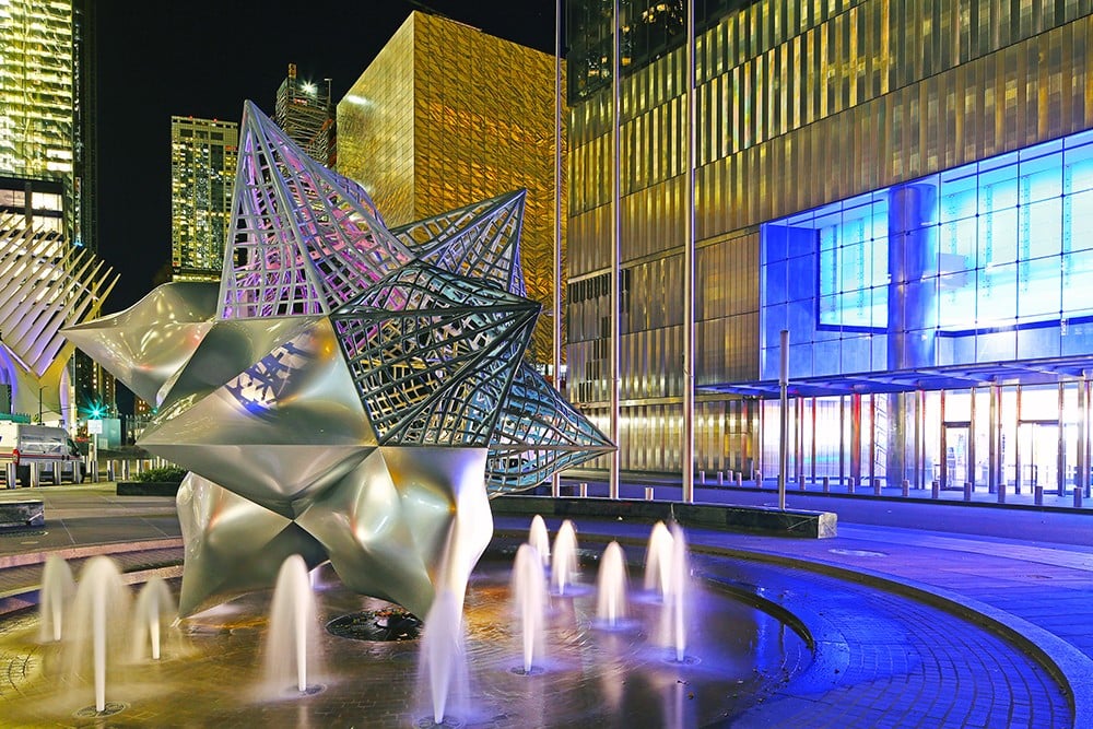 Frank Stella's Jasper's Split Star stainless steel sculpture illuminated at night beside a fountain at Silverstein Family Park at the World Trade Center.