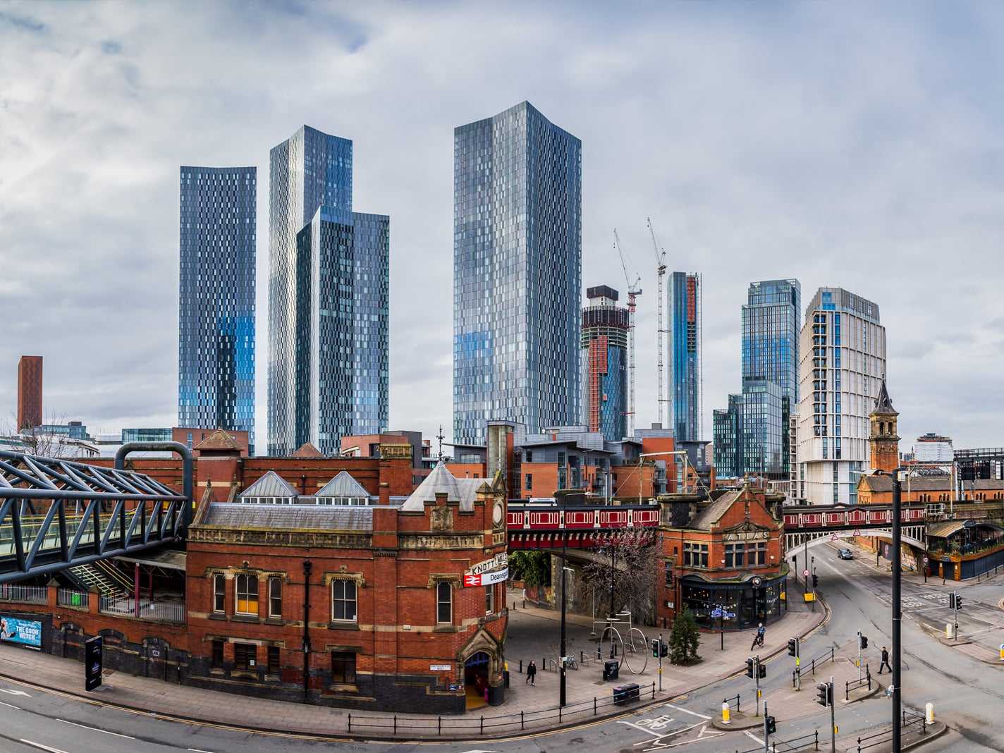 A wide-angle urban landscape of Manchester, UK, showcasing the striking contrast between the historic red-brick buildings of Castlefield and the modern glass skyscrapers of Deansgate Square