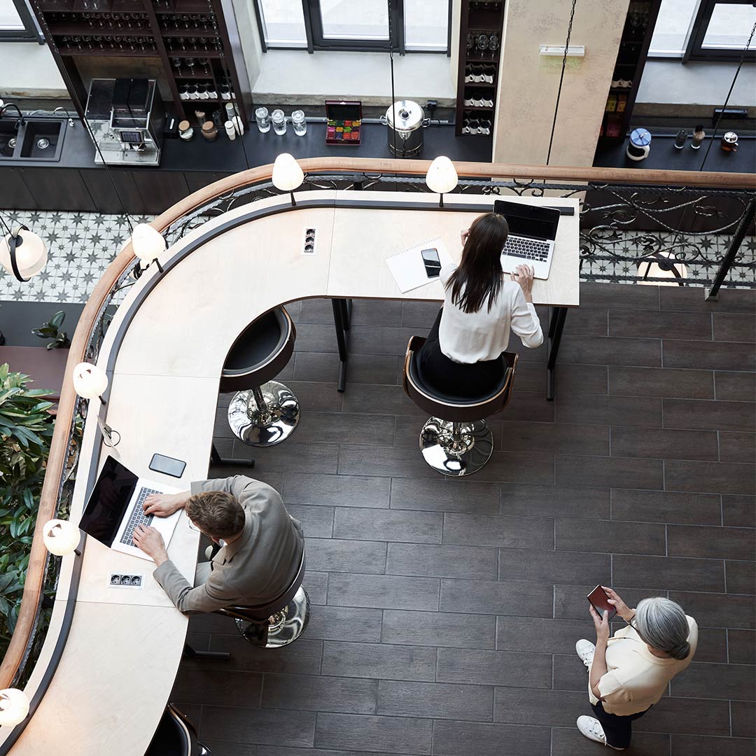 Bovenaanzicht van coworking ruimte met gebogen bar, mensen met laptops en koffie, lichte ramen, werkplek en horeca gecombineerd in open interieur.