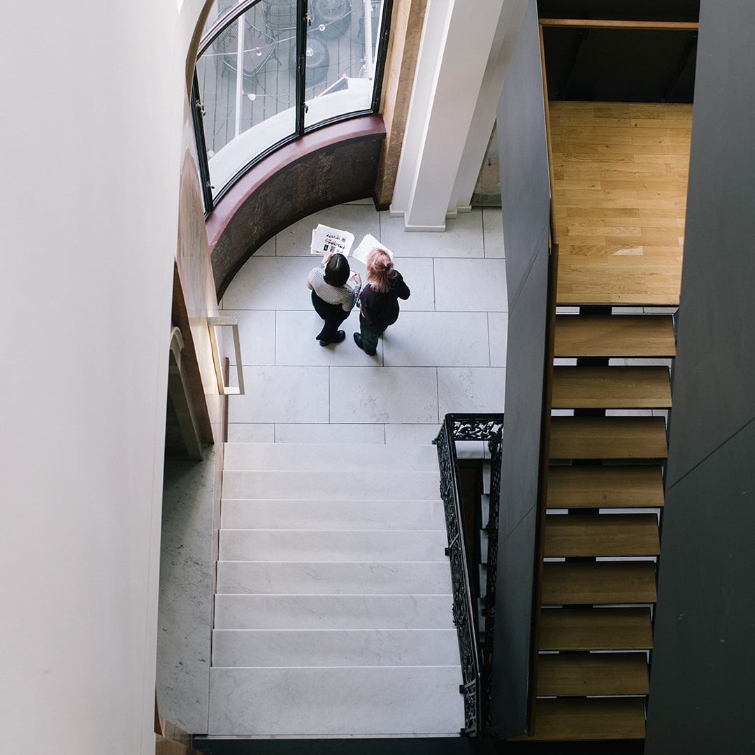 Overhead view of a modern interior atrium featuring marble flooring, a curved floor-to-ceiling window, two people reviewing documents, and adjacent staircases with white stone and wooden steps. 