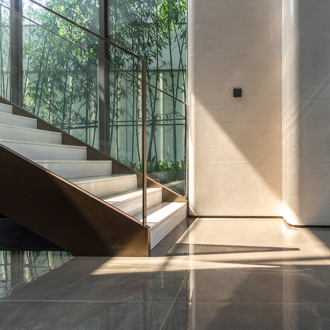 Sunlight streams across a polished modern interior, highlighting a glass-sided staircase and lush bamboo visible through floor-to-ceiling windows.