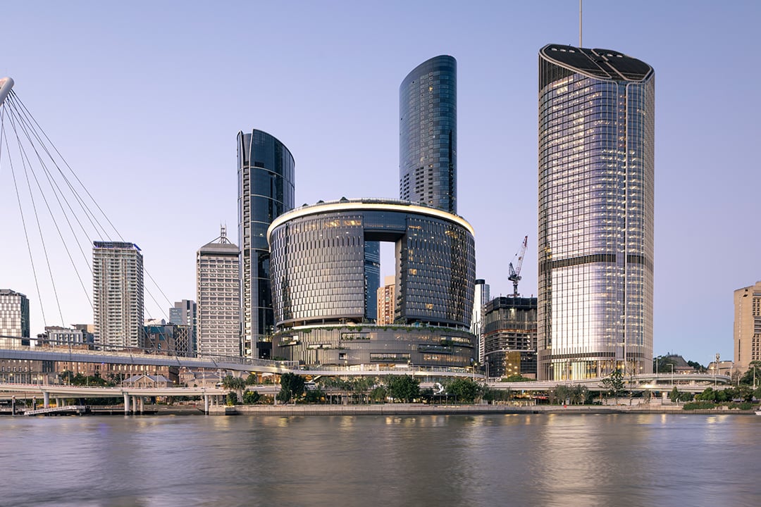 Riverfront city skyline with modern high-rise buildings, a pedestrian bridge and calm water in the foreground at dusk.