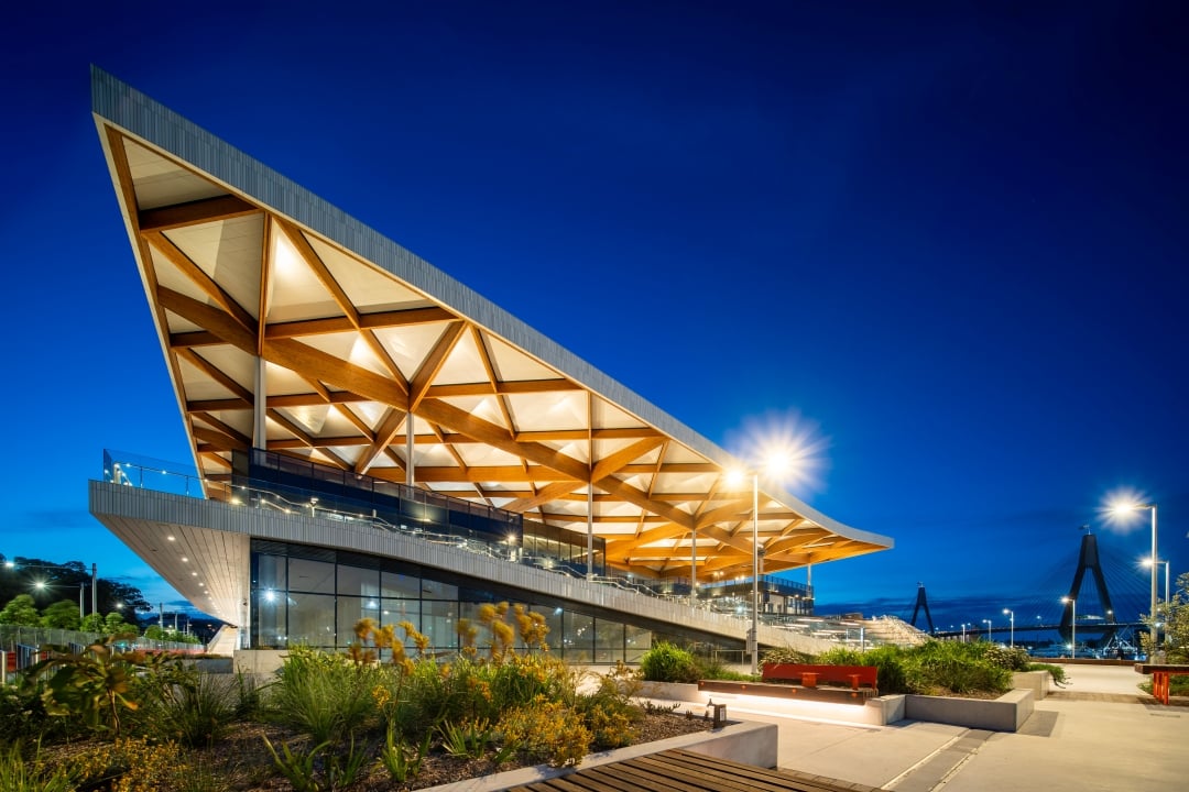 The new Sydney Fish Market building illuminated at night, featuring an angular timber roof, landscaped public areas and the Anzac Bridge in the background.