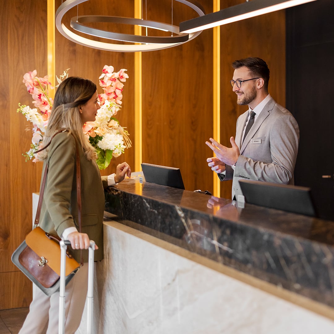 A hotel guest with luggage speaks with a receptionist at a modern reception desk in a warmly lit lobby. 