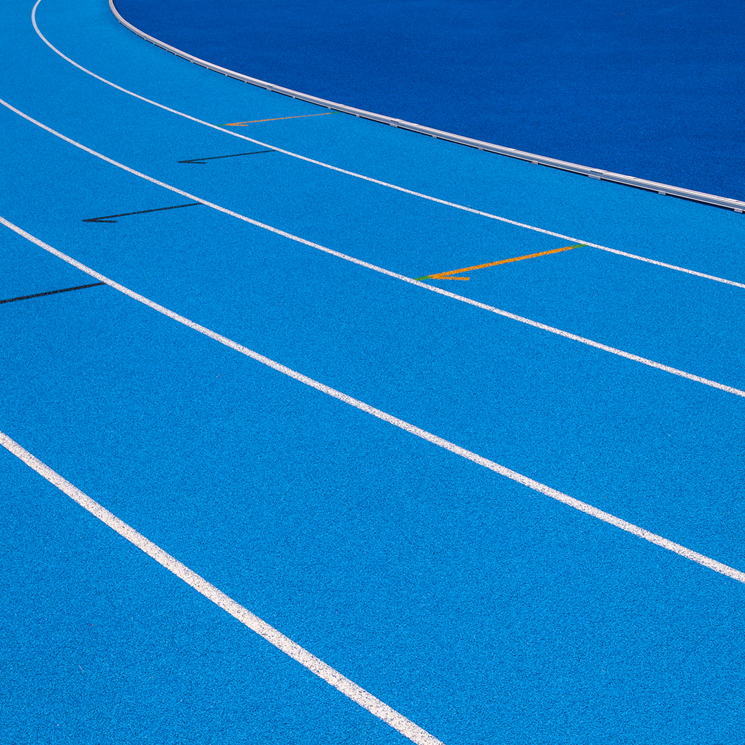 Curved lanes of a bright blue running track with white lane markings and directional arrows, captured from a close, angled perspective. 