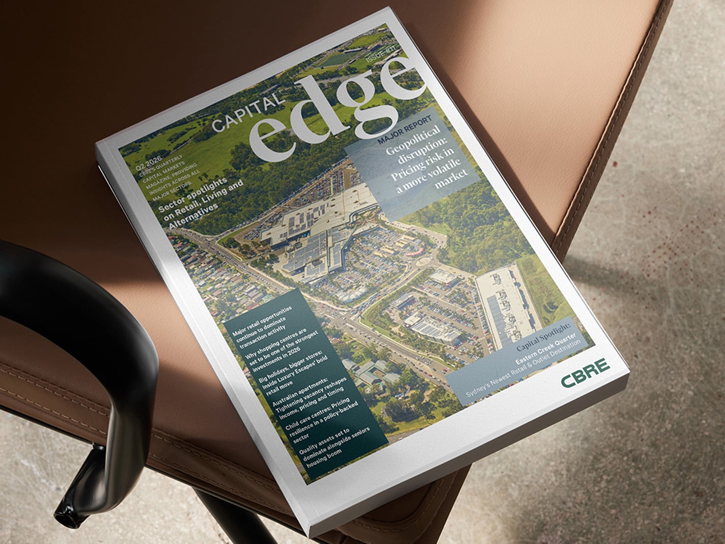A printed magazine titled “Capital Edge” rests on a brown leather chair, featuring an aerial view of a large commercial complex on the cover, with headlines about geopolitics, pricing risk, and market trends, and a CBRE logo in the bottom corner. 