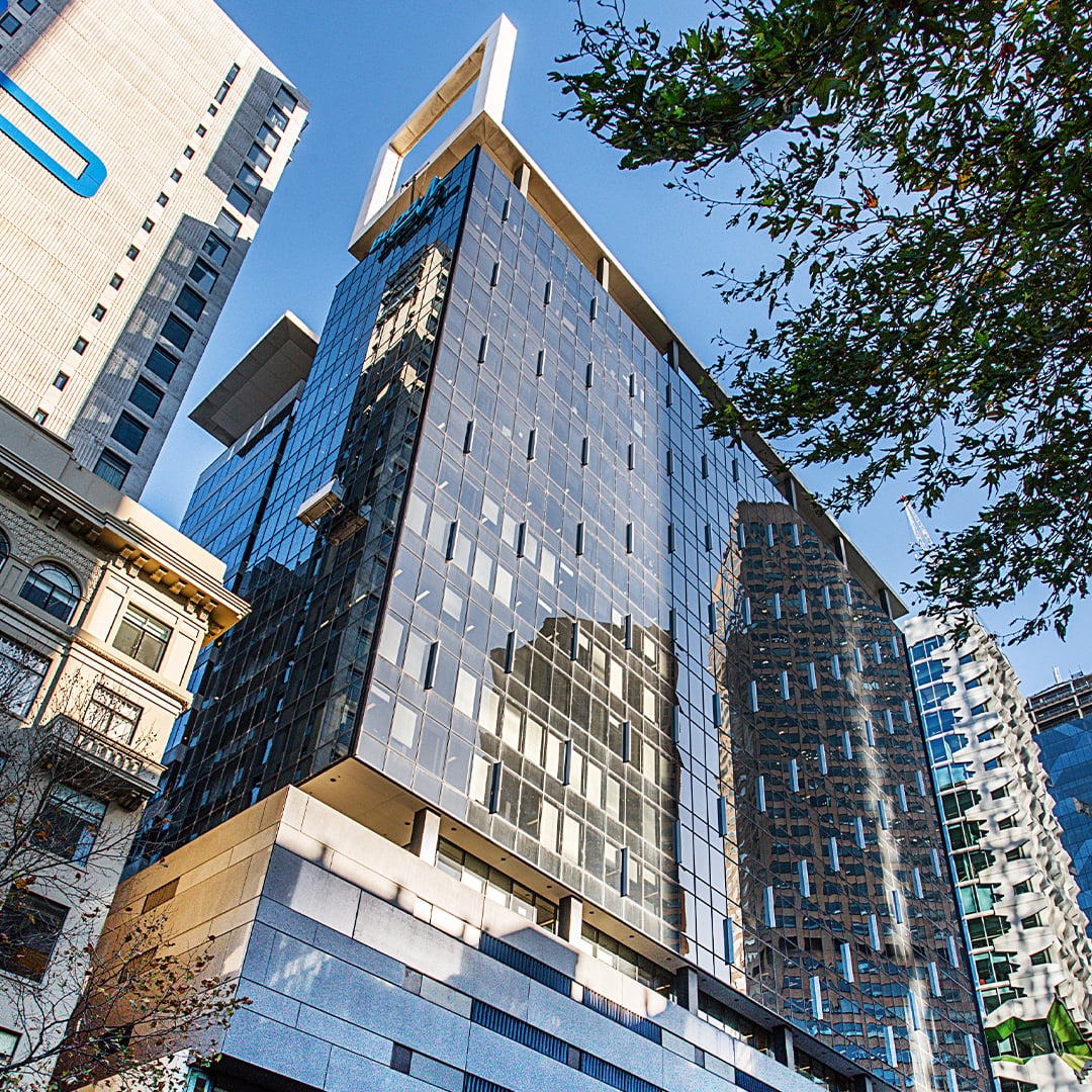 Modern glass-clad office tower reflecting surrounding city buildings, viewed from street level against a clear blue sky with tree branches framing the scene. 