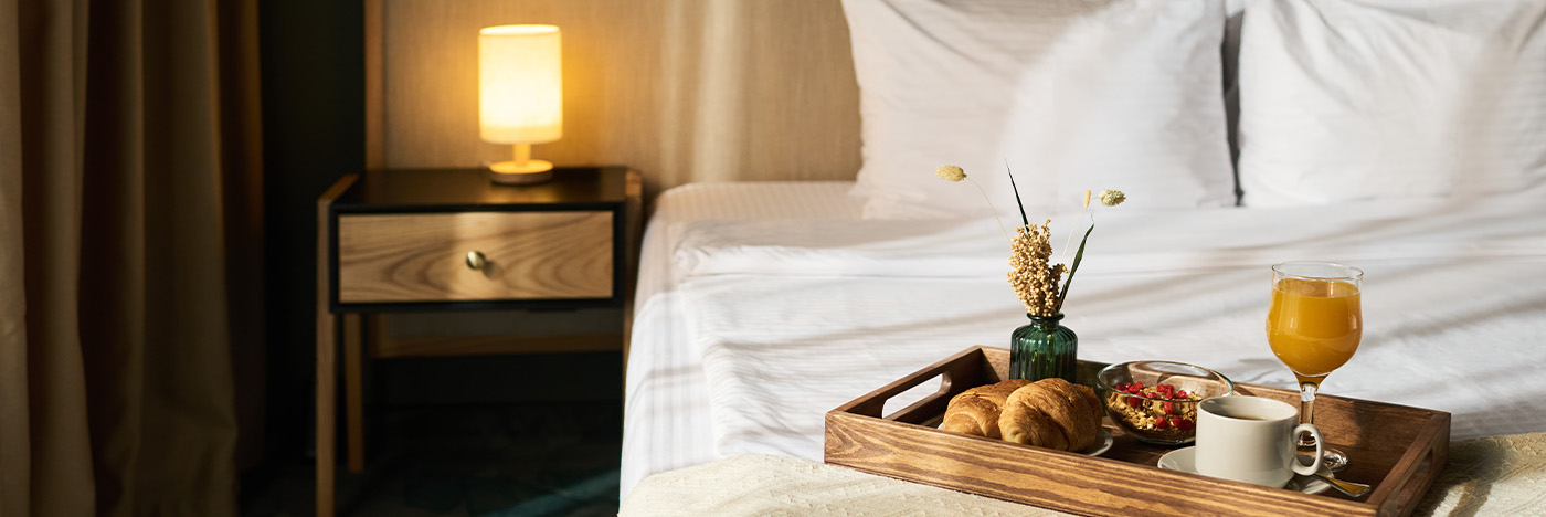 Hotel room with a breakfast tray on the bed, featuring pastries, coffee, and juice in a warmly lit setting.