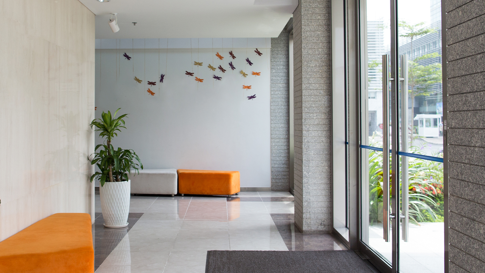 Modern building lobby with orange bench seating, a potted plant, decorative wall art, and floor-to-ceiling glass doors letting in natural light.
