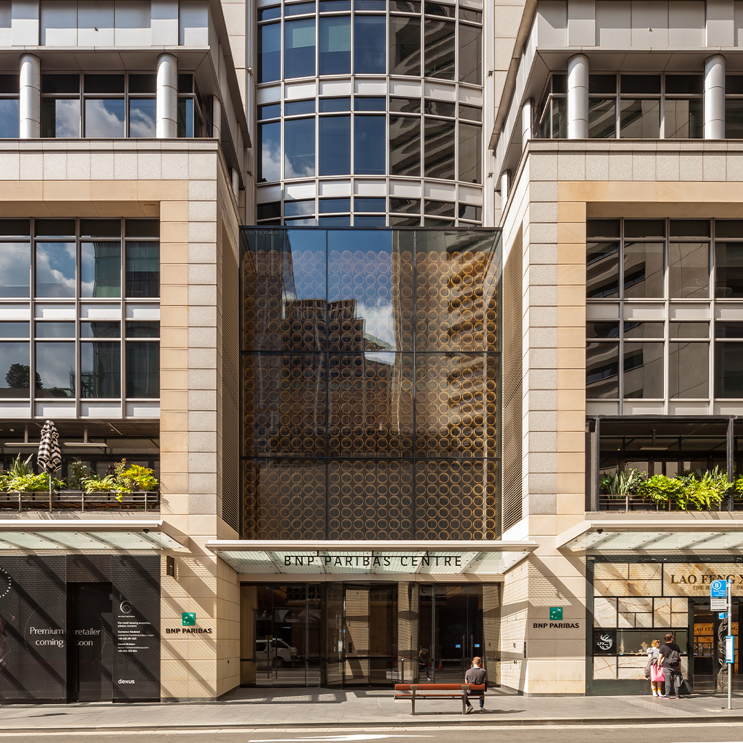 Street-level entrance to a contemporary office tower featuring a glazed lobby, stone-clad façade, retail frontage and landscaped terraces above the podium level.