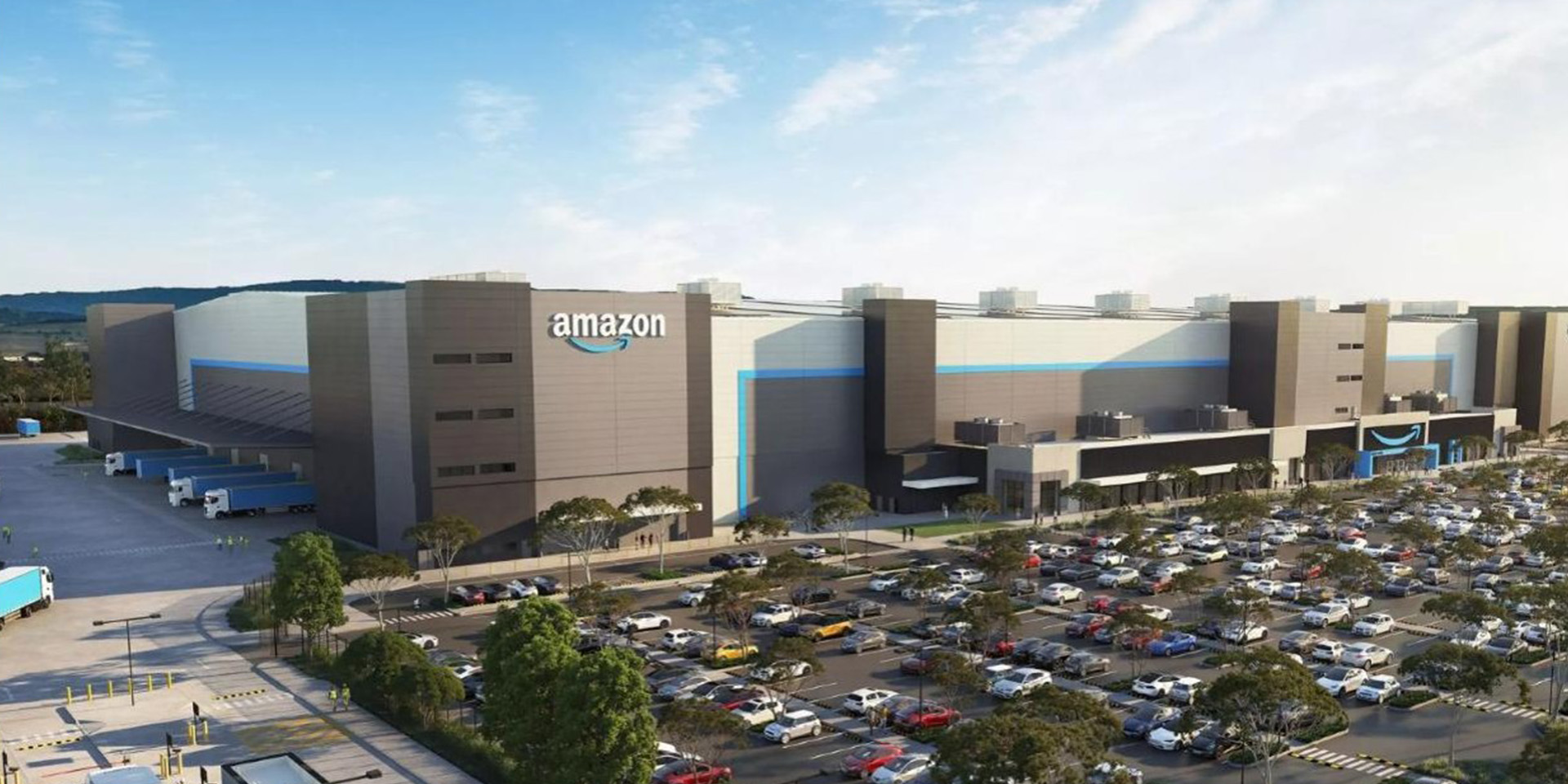 Amazon fulfillment center with a large warehouse building, truck loading bays, and a full parking lot filled with cars, viewed from an elevated angle under a clear sky.