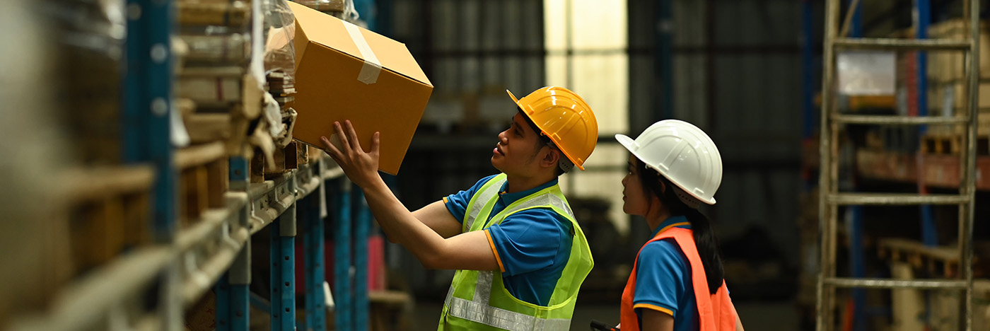 Warehouse workers wearing safety helmets and high-visibility vests place a cardboard box onto a storage shelf inside a large industrial warehouse aisle lined with stocked racks.