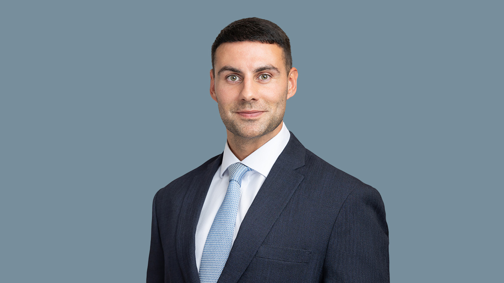 Professional headshot of Sam Saghir Pashley in a dark business suit and light blue tie against a plain blue-grey background.