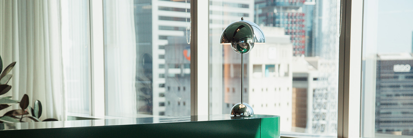 Modern office interior with a sleek countertop and chrome table lamp positioned by large windows overlooking a city skyline with high-rise buildings.