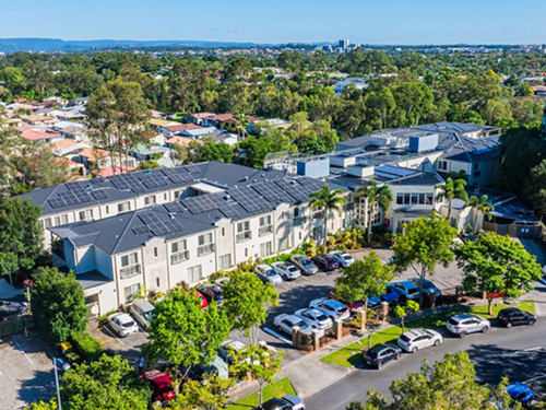 Aerial view of a multi-storey aged care or residential facility with solar panels, landscaped grounds, and parking areas within a suburban neighbourhood. 