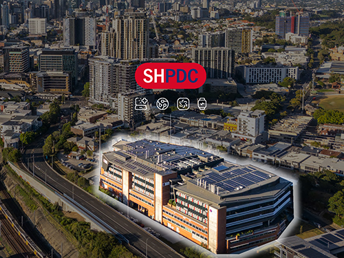 Aerial view of a modern multi-storey commercial office building with rooftop solar panels, highlighted within a dense inner-city precinct surrounded by high-rise residential and commercial developments. 