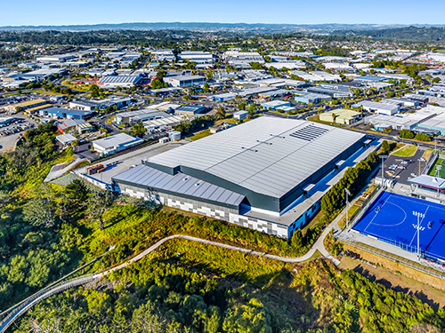 Aerial view of a large industrial warehouse complex surrounded by commercial buildings, roads, and green space on the edge of an urban area. 